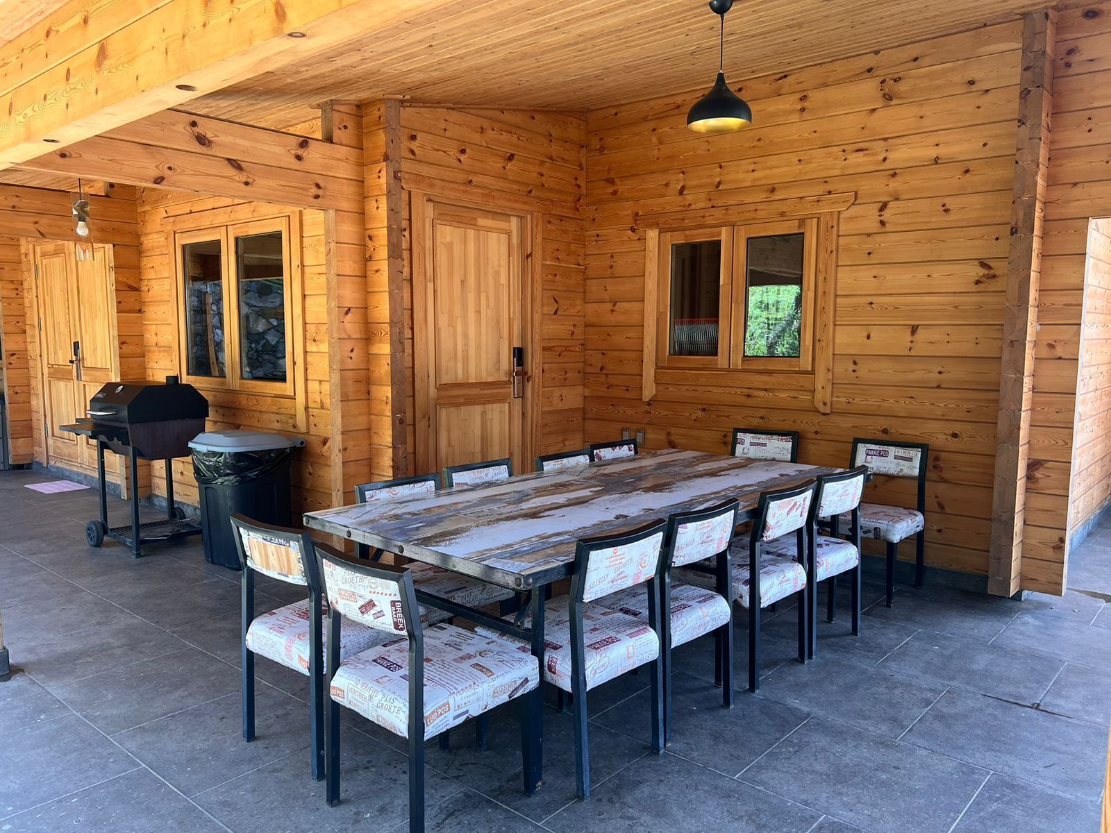 Una cabaña de madera con una gran mesa de comedor y sillas, una parrilla en el exterior y un cartel que anuncia venta de pasteles.