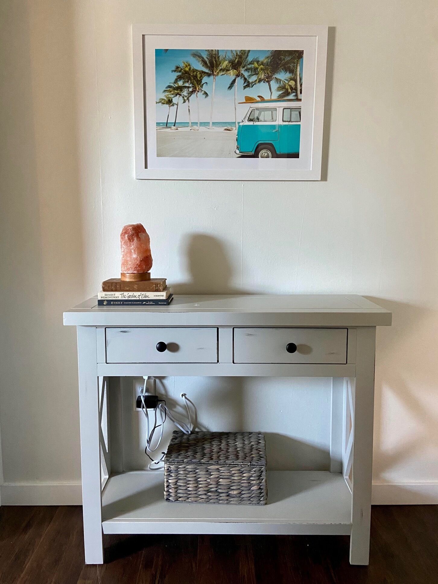 A white console table with a framed picture above it, a salt lamp, and a woven basket below.