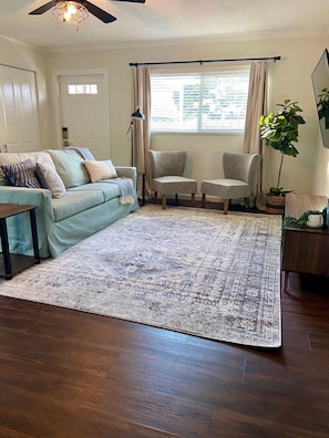 A living room with a light blue sofa, two armchairs, a coffee table, a patterned rug, a potted plant, and a ceiling fan.