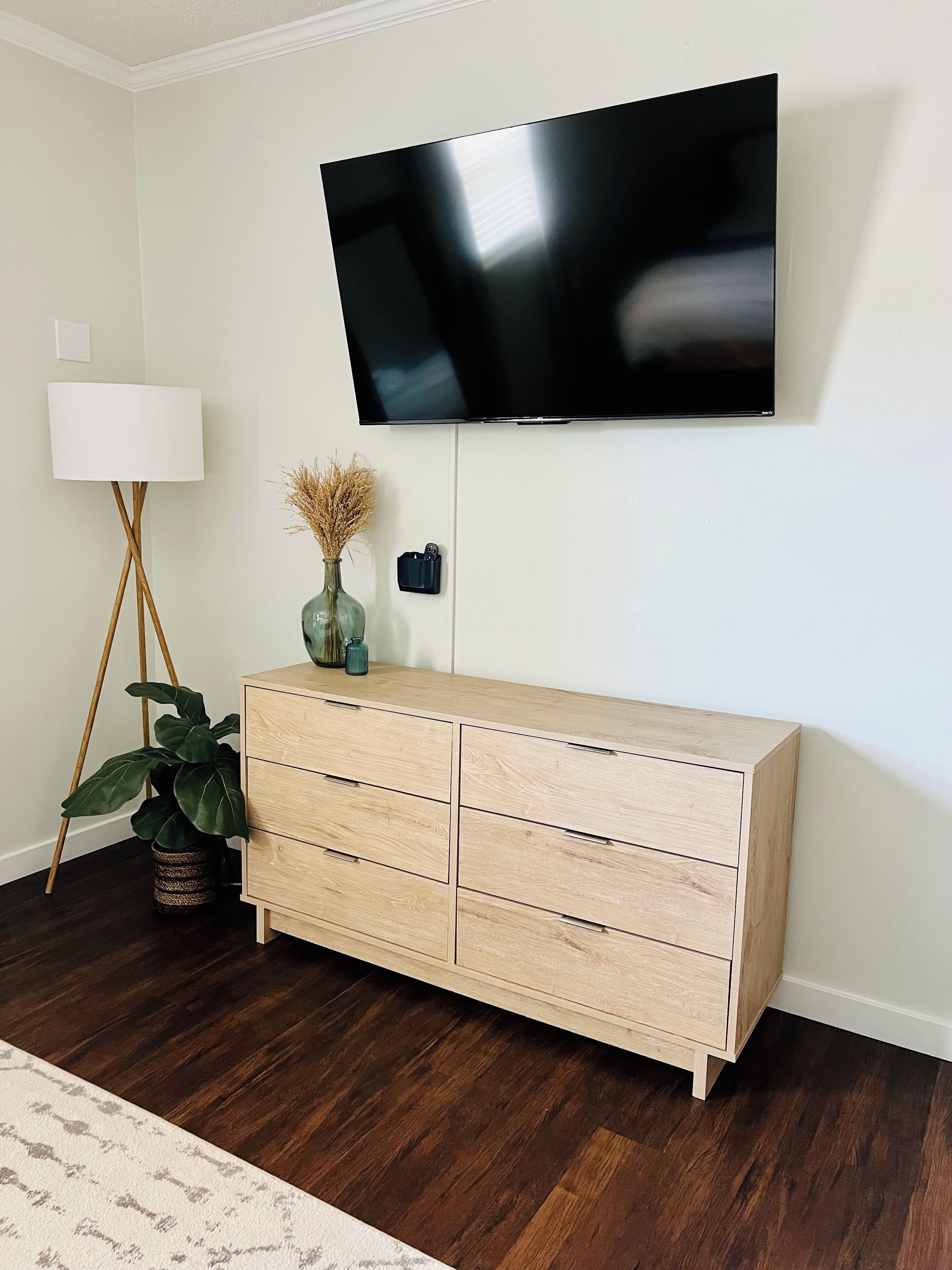 A room with a wooden cabinet, a flat-screen TV mounted on the wall, a floor lamp, and a potted plant.