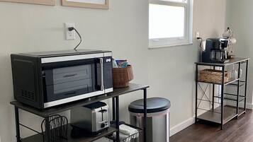 Une cuisine équipée d’un four à micro-ondes, d’un grille-pain et d’une machine à café, le tout posé sur un support en métal noir. Des tableaux encadrés ornent le mur, et une fenêtre offre une vue sur l’extérieur.