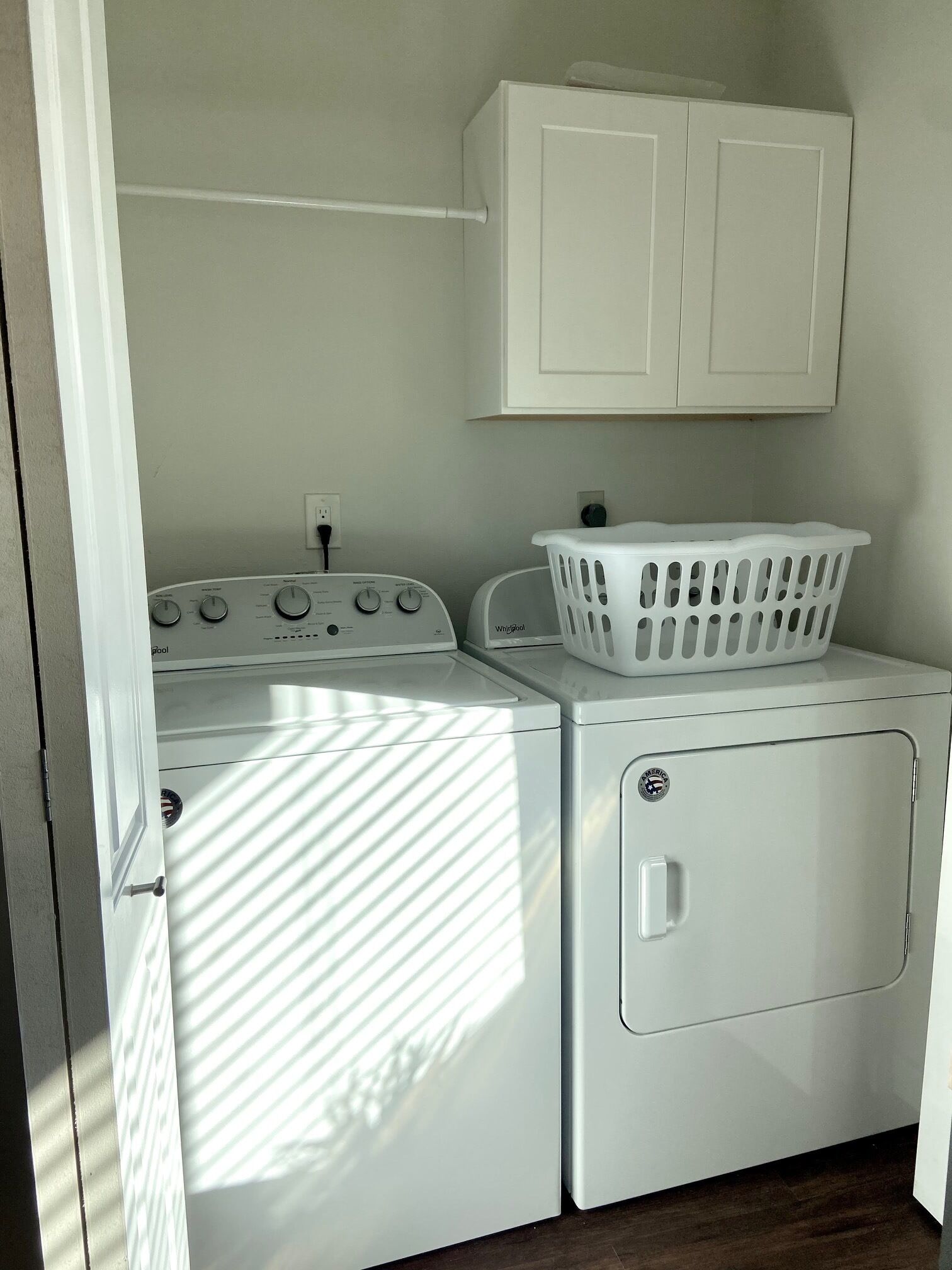 A laundry area with a front-loading washer and dryer, a white laundry basket, and a white cabinet above the dryer.