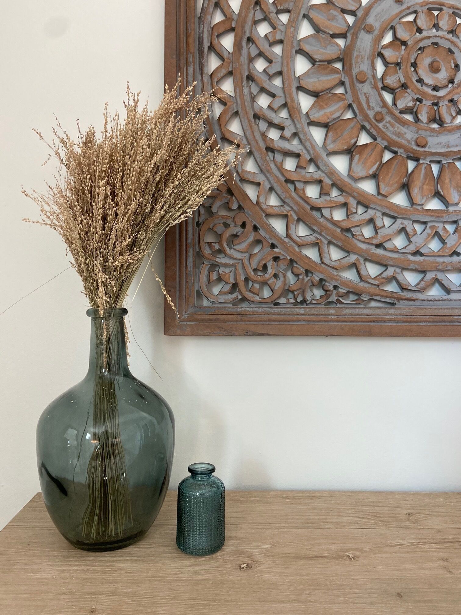 A vase with dried plants on a wooden table, a decorative metal piece on the wall, and a small green bottle beside the vase.