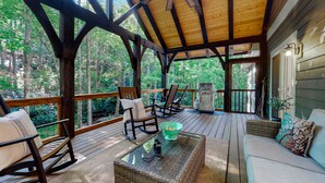 Une terrasse couverte avec un plancher en bois, des fauteuils à bascule, une table à plateau en verre et un barbecue.