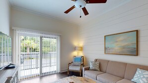 A living room with a beige sofa, a wooden coffee table, a ceiling fan, a painting, and a view through a sliding glass door. - King one-bedroom condo w/ pool in the SSI Village area Unit 2205 (Saint Simons Island)