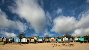 A row of beach huts with a sandy beach and a partly cloudy sky. - Penmorfa - Sublime Abersoch retreat with roof terrace, 80m from beach, great for surfing (Abersoch)