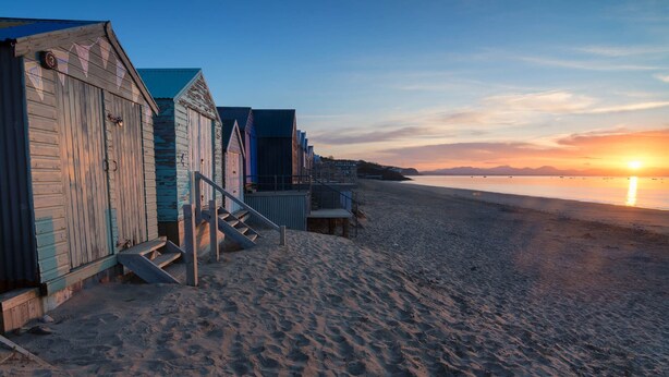 A row of beach huts with steps leading up to them, positioned along a sandy beach with a sunset in the background. - Penmorfa - Sublime Abersoch retreat with roof terrace, 80m from beach, great for surfing (Abersoch)