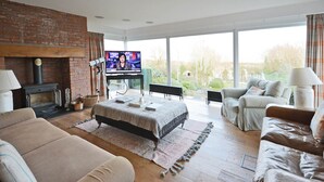 A spacious living room with a fireplace, a television, and a large window offering a view of the outdoors. - Penmorfa - Sublime Abersoch retreat with roof terrace, 80m from beach, great for surfing (Abersoch)