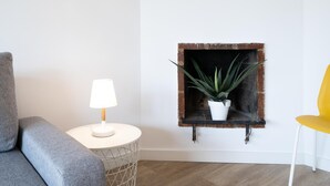 Ett modernt vardagsrum med en grå soffa, ett vitt sidobord med en lampa, en rustik hylla med en växt och en gul stol.