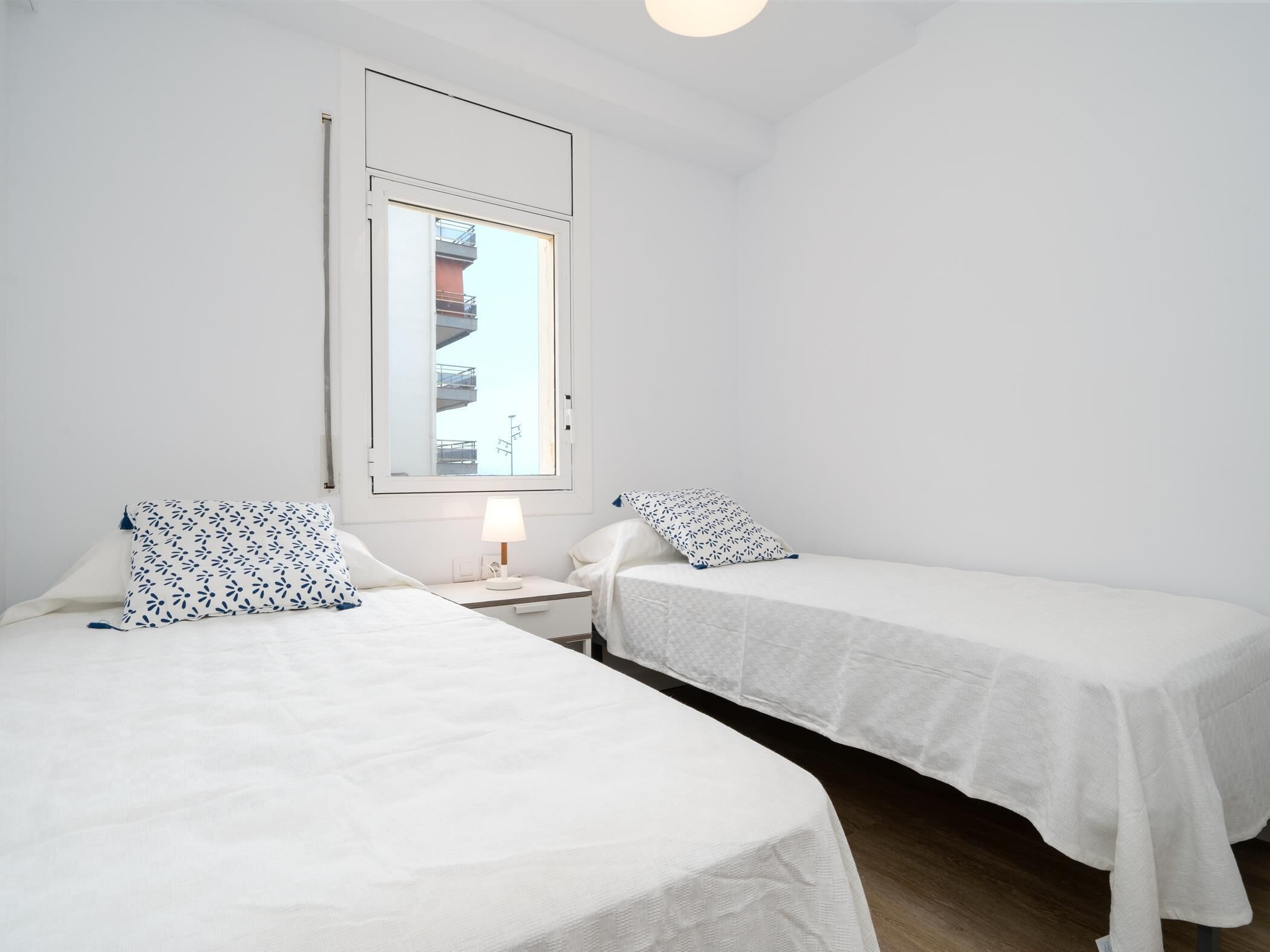 Una habitación pequeña y limpia con dos camas individuales, una mesita de noche con lámpara y ventana con vistas a edificios.
