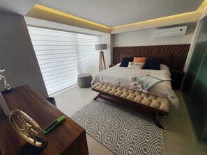 A modern bedroom with a large bed, a desk, and a window with blinds.