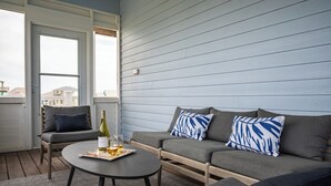Ein Balkon mit einer grauen Couch, zwei blau-weißen Kissen, einem runden Tisch mit einer Weinflasche und Gläsern, sowie einem gemusterten Teppich.