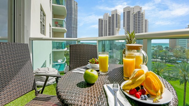 A balcony with wicker furniture, a dining table with breakfast items, and a view of tall buildings and greenery. - OCEAN RESERVE LUXURIOUS CONDO 2MIN AWAY FROM BEACH (Sunny Isles Beach)
