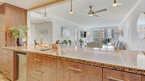 A modern kitchen with granite countertops, wooden cabinets, and a central island. There are ceiling fans, pendant lights, and a large window offering a view of the city. - OCEAN RESERVE LUXURIOUS CONDO 2MIN AWAY FROM BEACH (Sunny Isles Beach)