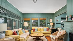 A cozy room with a green wall, a yellow sofa, a round wooden coffee table, wicker chairs, and a fireplace.