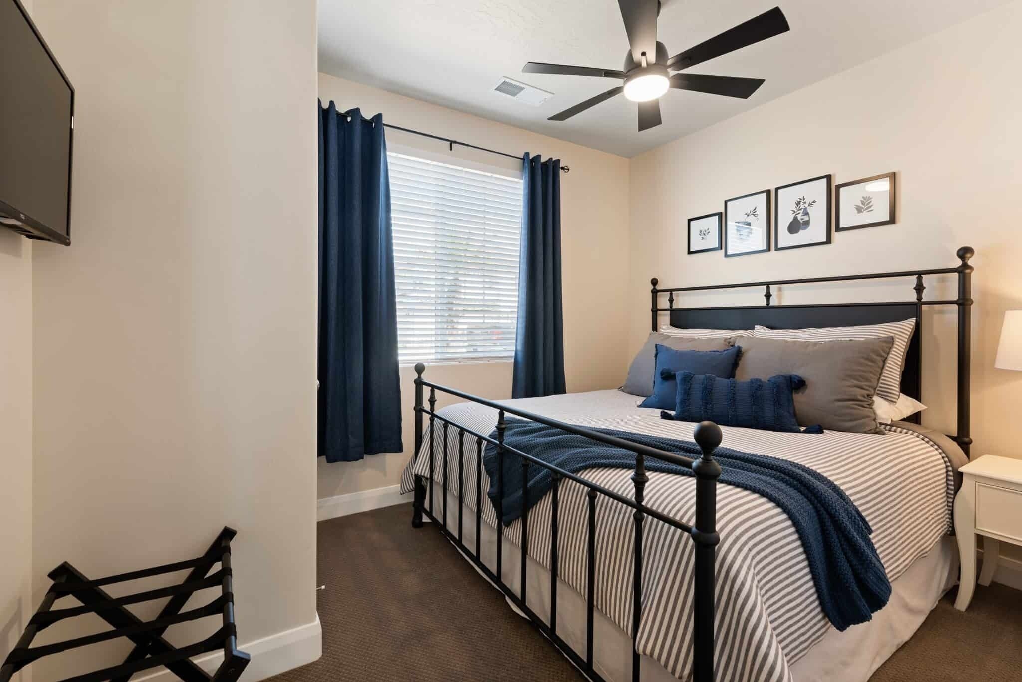 A bedroom with a bed, a ceiling fan, a window with curtains, and framed pictures on the wall.