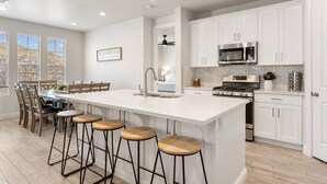 A modern kitchen with white cabinets, a large island with stools, a microwave, stove, and a view of the outdoors through large windows. - The Gathering Place at Escondido by RedAwning (Washington)