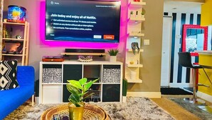 Una sala de estar con un televisor de pantalla plana en la pared, un sofá azul, una mesa de centro con una planta y una estantería decorativa.