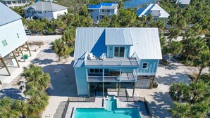 Uma casa à beira-mar de dois andares, com piscina, rodeada por palmeiras e outras casas de praia.