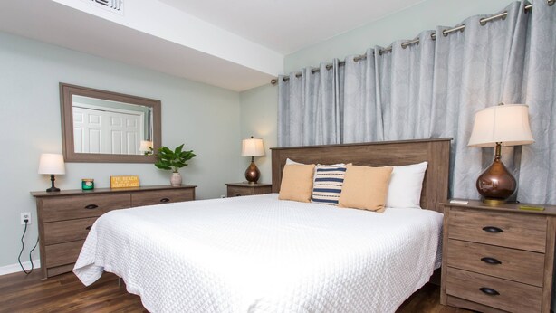A bedroom with a large bed, two bedside tables with lamps, a dresser, a mirror, and a potted plant. - Next to Kayak Park |1st Floor Condo |Short Walk to Beach |Beach Cart, 2 Chairs & Umbrella |Dogs Ok (Fort Walton Beach)