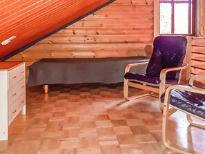 A log cabin interior with a wooden floor, a bed with a grey cover, and two chairs.