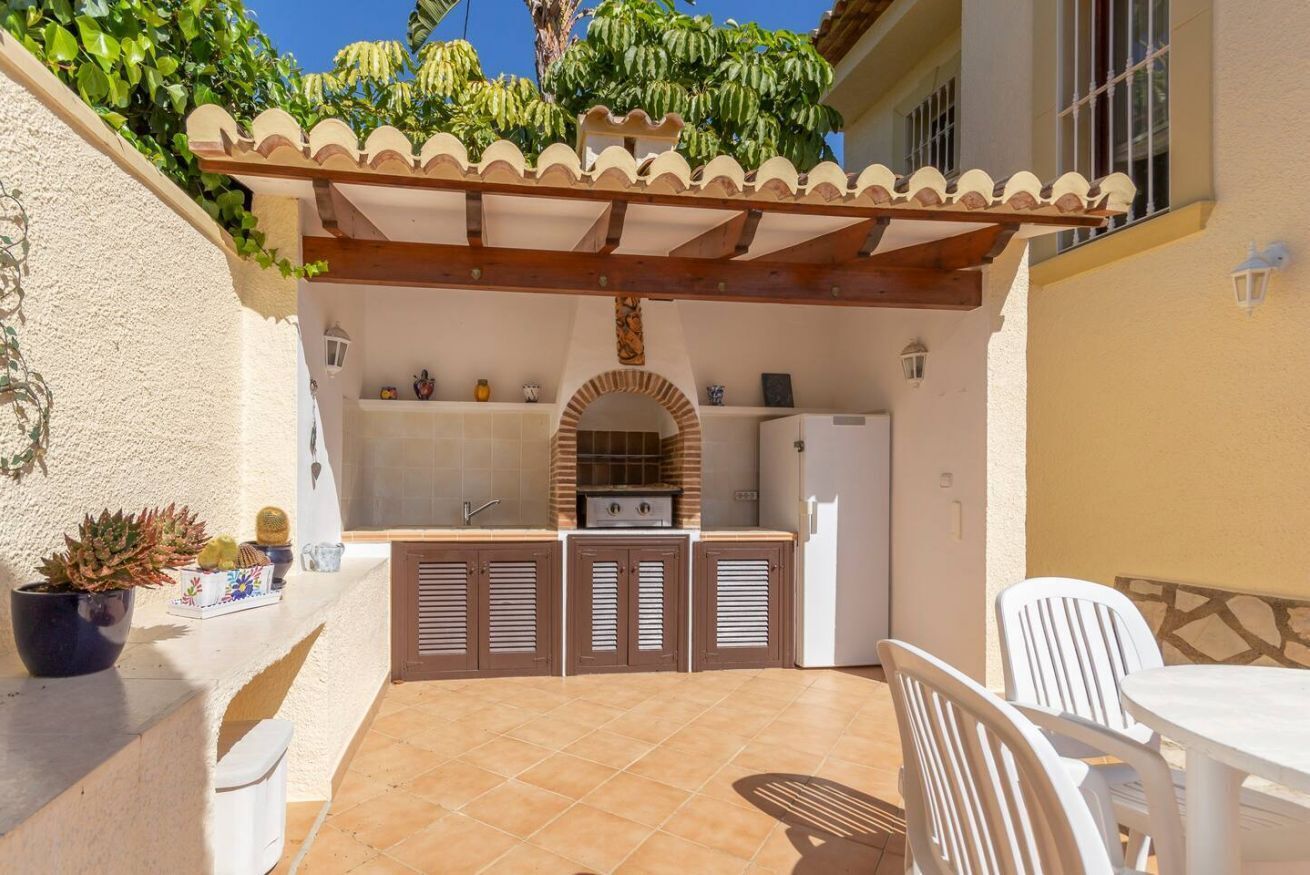 A patio with a built-in kitchen, including a grill, white chairs, and a tiled floor.