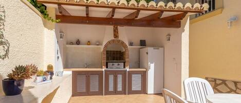 A patio with a built-in kitchen, including a grill, white chairs, and a tiled floor.
