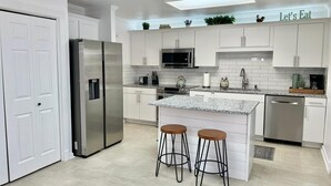 Una cocina moderna con armarios blancos, un refrigerador de acero inoxidable, un microondas y una cafetera. Hay dos taburetes en la isla.