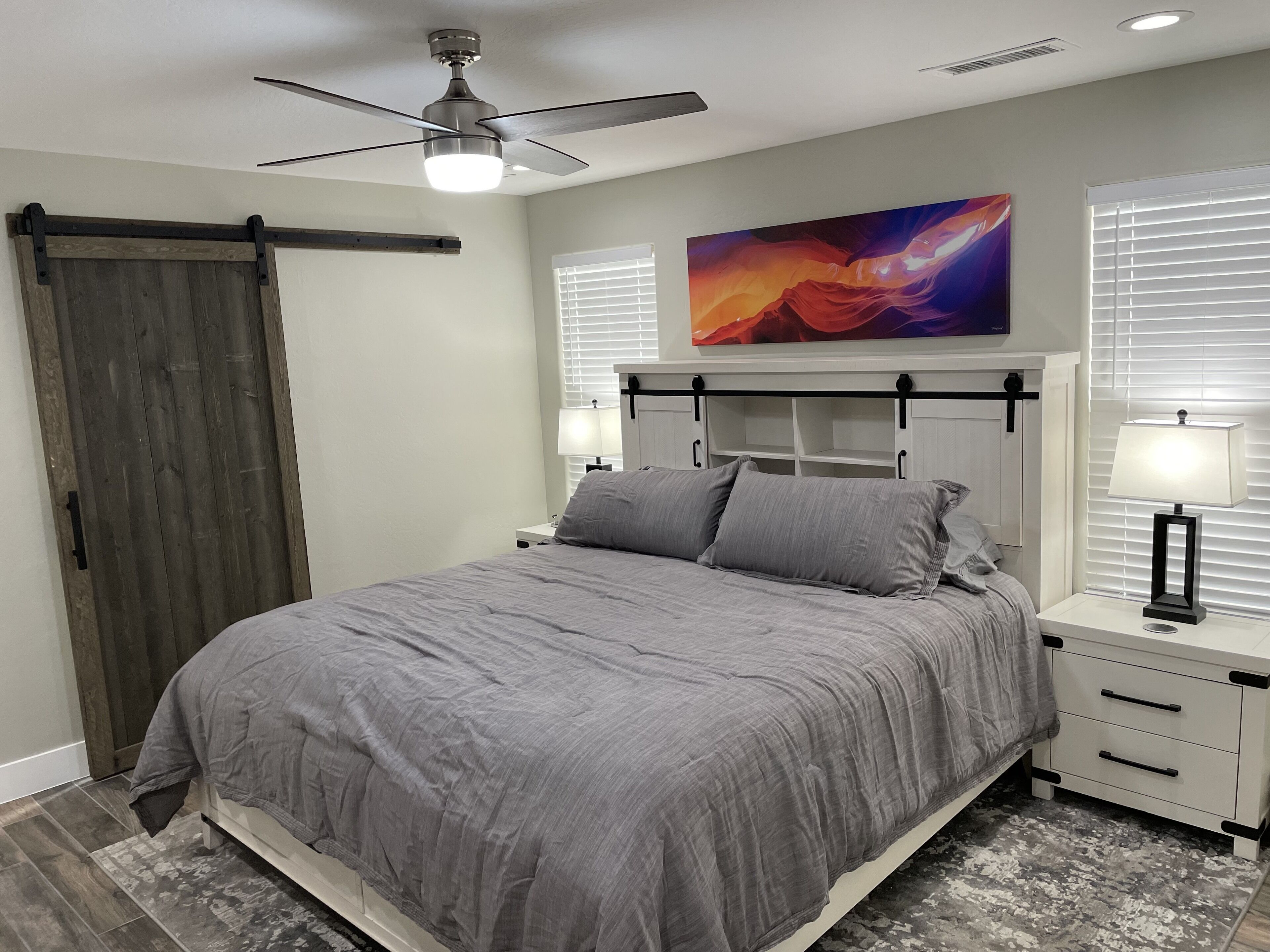 Un dormitorio con una cama, un ventilador de techo, una mesita de noche, una lámpara y un cuadro en la pared.