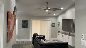 Un salon moderne avec un canapé d’angle noir, un ventilateur de plafond, un éclairage encastré, un téléviseur à écran plat et un coin repas avec une petite table et des chaises.