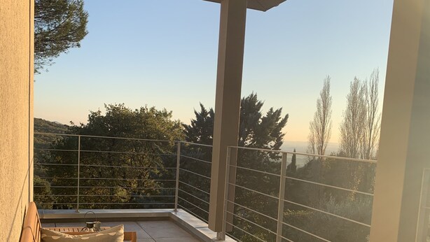 Eine hölzerne Bank mit Kissen auf einem Balkon mit Metallgeländer, mit Blick auf eine hügelige Landschaft.