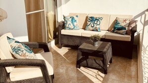 Une terrasse ensoleillée, meublée avec des meubles en osier, notamment un canapé et des fauteuils, ainsi qu’une petite table agrémentée d’une plante.