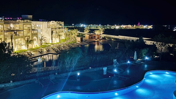 Vue nocturne d’un hôtel doté d’une grande piscine, de palmiers et d’un bâtiment bien éclairé, orné de nombreux balcons.