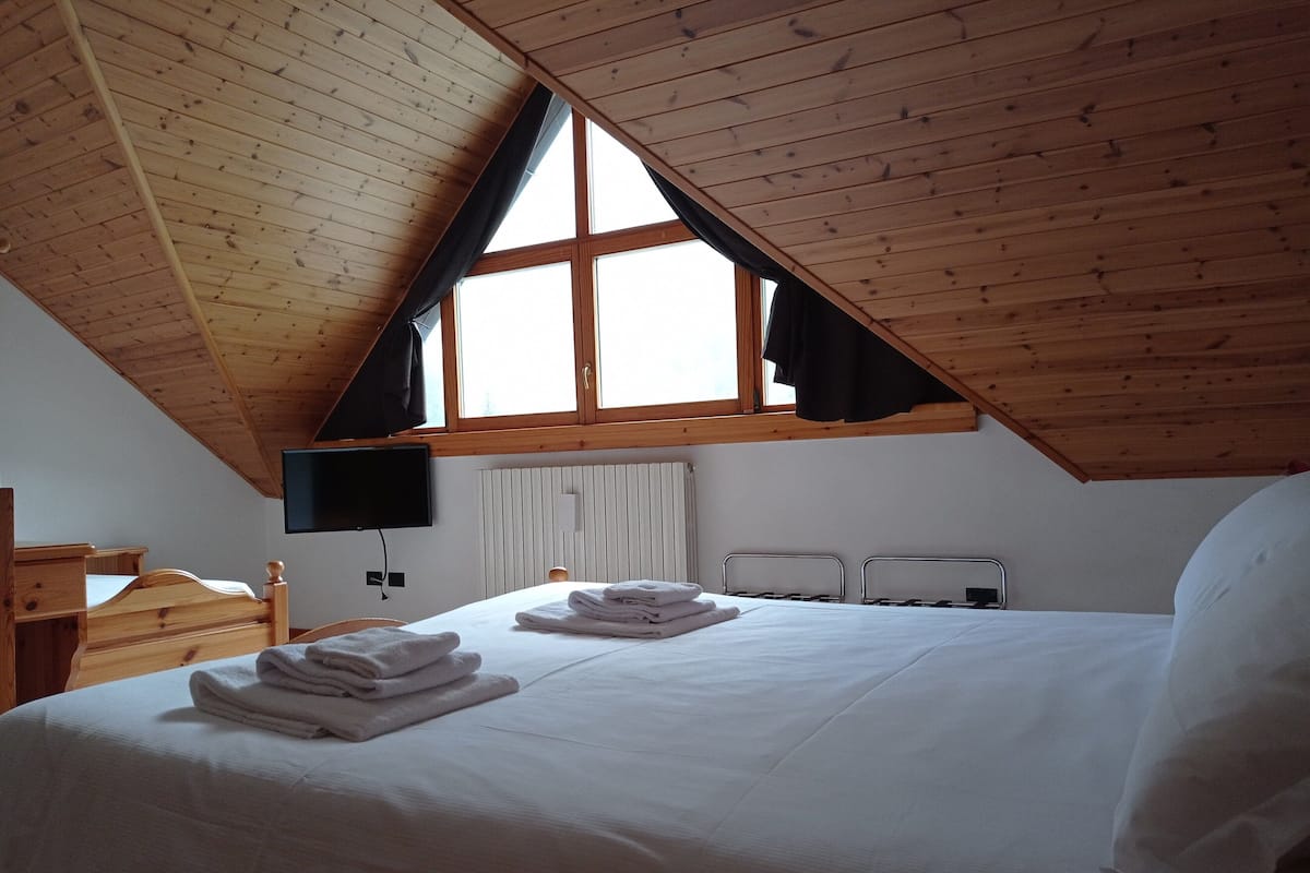 A bedroom with a sloped ceiling, a bed with white linens, a flat-screen TV mounted on the wall, and a window with curtains.