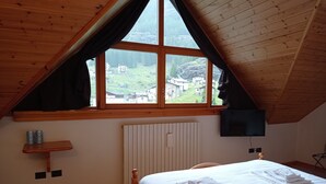 A bedroom with a sloped ceiling, a large window with a view of a village, a bed with white linens, a shelf with bottles, and a wall-mounted TV. - Hotel Fior di Roccia Valmalenco (Lanzada)
