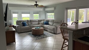 A spacious living room with a large sectional sofa, a dining area with a wooden table and chairs, and a kitchen with a white countertop.