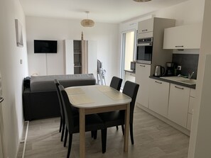 En kompakt opholdsstue med spisebord, stole, sofa, tekøkken og et tv ophængt på væggen.