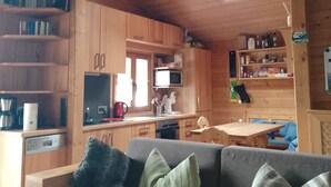 Ein gemütliches Blockhaushaus-Interieur mit Holzbalkendecke, einer Couch mit Kissen, einem Esstisch und einer Küchenzeile mit Holzschränken und Geräten.