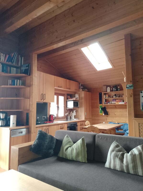 A cozy cabin interior with a wooden ceiling, a sofa with cushions, a dining table, and a kitchen area with wooden cabinets and appliances.