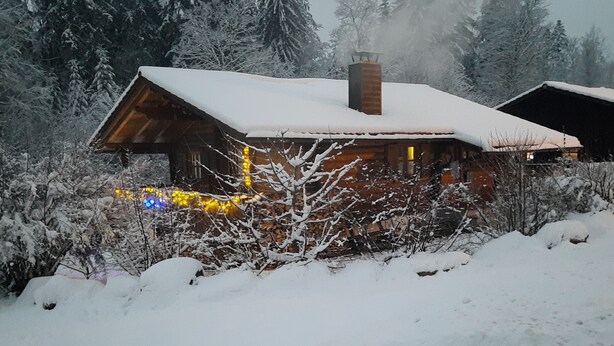 Eine Holzhütte mit schneebedecktem Dach und Schornstein, umgeben von schneebedeckten Bäumen und Sträuchern.