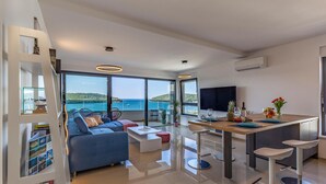 A modern living room with a sofa, coffee table, and a dining area with a table set for two. There is a large window offering a view of the sea and a balcony. - 2 bedroom beautiful apartment in Banjole (Banjole)