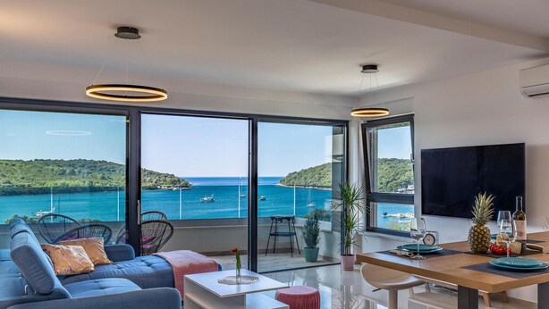 A modern living room with a large window overlooking a coastal view, a dining area with a wooden table, and a flat-screen TV mounted on the wall. - 2 bedroom beautiful apartment in Banjole (Banjole)