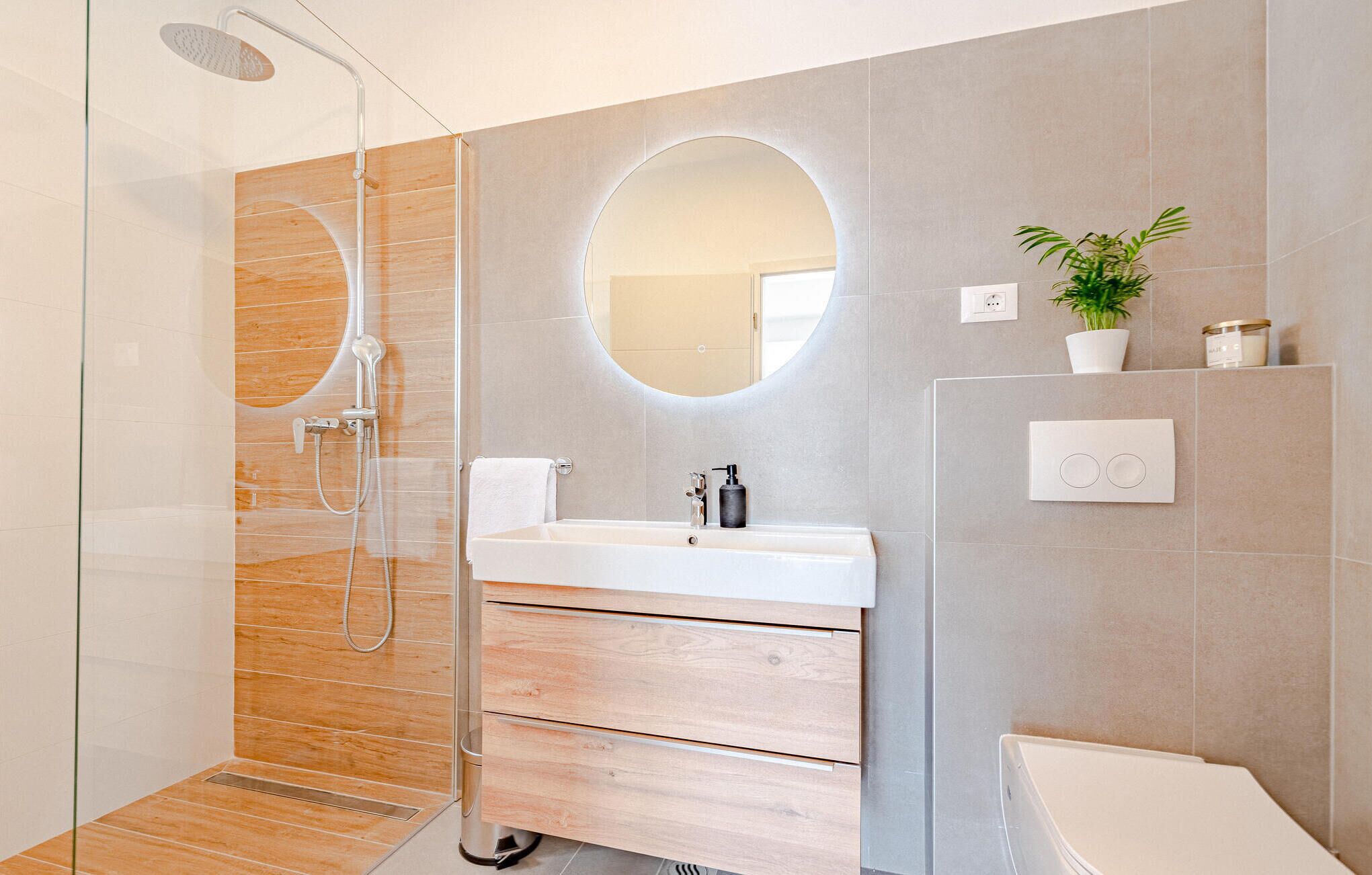 Een moderne badkamer met een douchecabine van glas, een houten wastafelmeubel, een grote spiegel en een potplant.