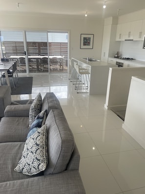 A modern living room with a grey sofa, a kitchen area with white cabinets, and a dining area with a table and chairs. - 4 Bedroom Beach House with Sea Views. Now with wifi and foxtel. (Kingston)