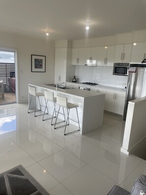A modern kitchen with white cabinetry, a central island with bar stools, a microwave, and a refrigerator. - 4 Bedroom Beach House with Sea Views. Now with wifi and foxtel. (Kingston)