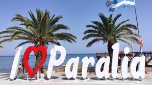 Ein Strand mit Palmen und einem Schild mit der Aufschrift 'I ♥ Paralia', im Hintergrund weht eine griechische Flagge.