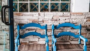 Zwei blaue Stühle mit gewobenen Sitzflächen und Lehnen stehen vor einer Steinwand mit einem Fenster und bläulich schimmernden Glasscheiben.