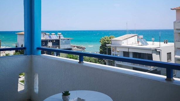 Ein Balkon mit Tisch und Stühlen, der Blick auf die Küste mit Gebäuden und dem Meer freigibt.