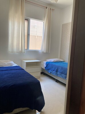 A bedroom with two beds, a window with curtains, a white desk, and a patterned bedspread.