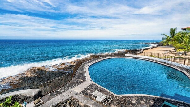 A pool with a curved edge, surrounded by a stone wall and a railing, overlooking the ocean.